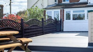 Ein Patio im Freien mit einem Holztisch und Bänken, einem schwarzen Metalltor und einem weißen Gebäude mit Fenstern im Hintergrund.
