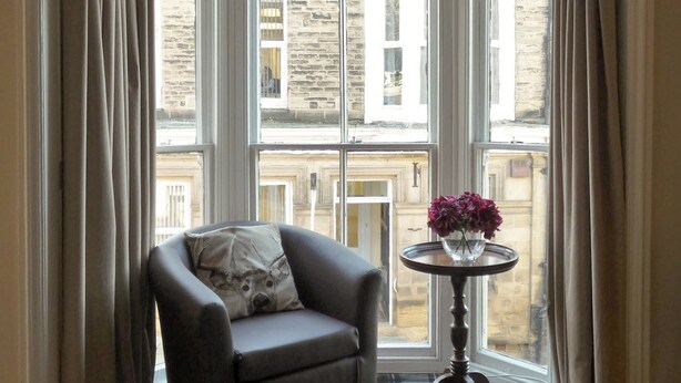 Une pièce d’angle avec un fauteuil sombre, une petite table ronde ornée d’un vase de fleurs et une vue sur un bâtiment en pierre, visible à travers de grandes fenêtres.