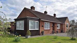 Une maison à deux étages, construite en briques, avec un toit en tuiles, plusieurs cheminées et une pelouse bien entretenue où poussent des jonquilles.
