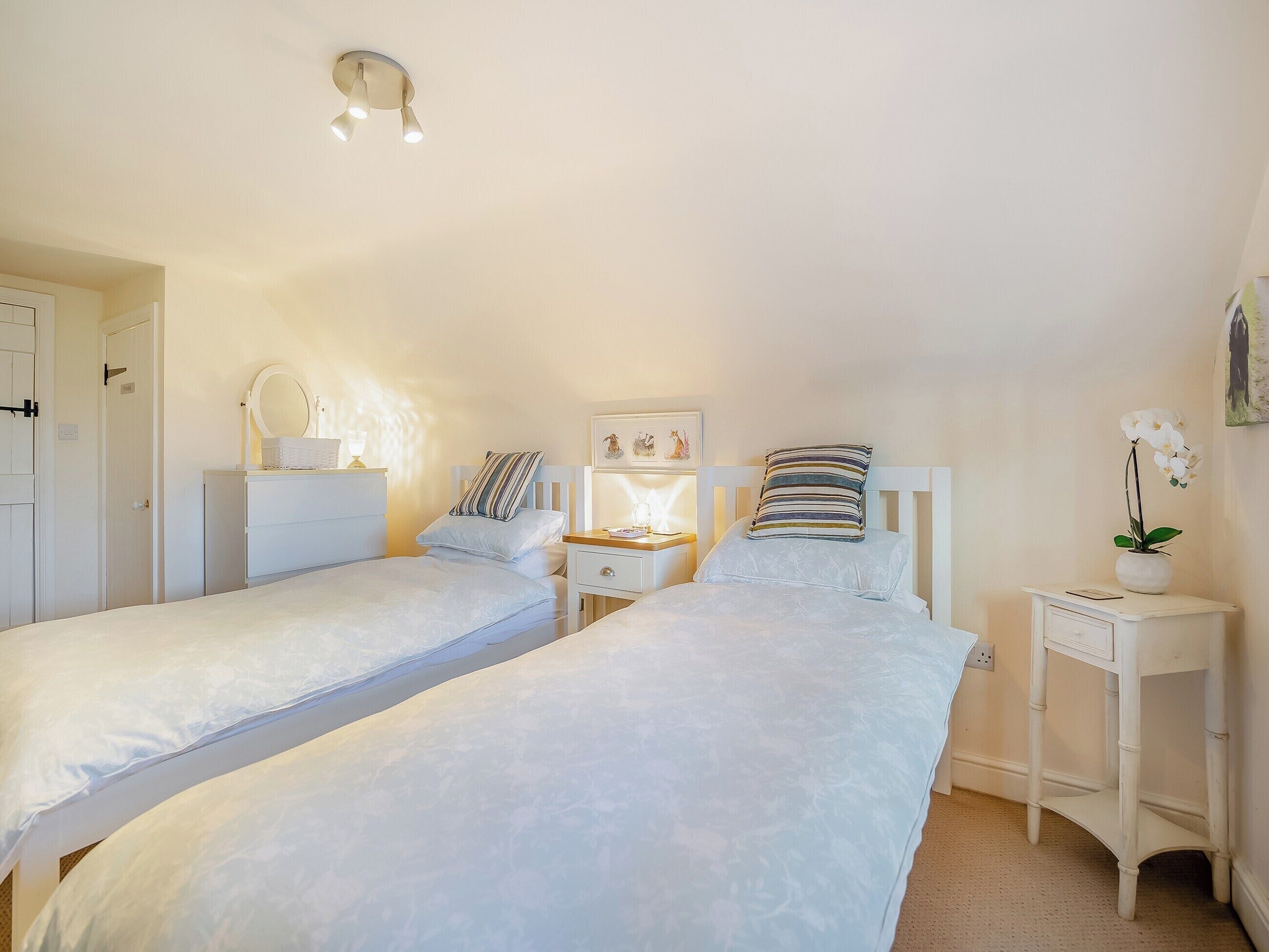 A bedroom with two single beds, a bedside table with a lamp, a small dresser, and a wall-mounted light fixture.
