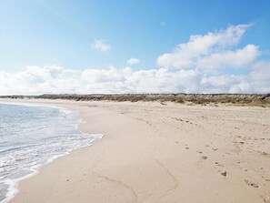 A wide sandy beach with gentle waves, a clear sky, and dunes in the background.