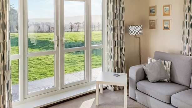 A living room with a sofa, a small table, and a floor lamp. There is a large window with curtains, and a view of a green field and trees. - 2 bedroom accommodation in Dulas, Anglesey (Dulas, Anglesey)