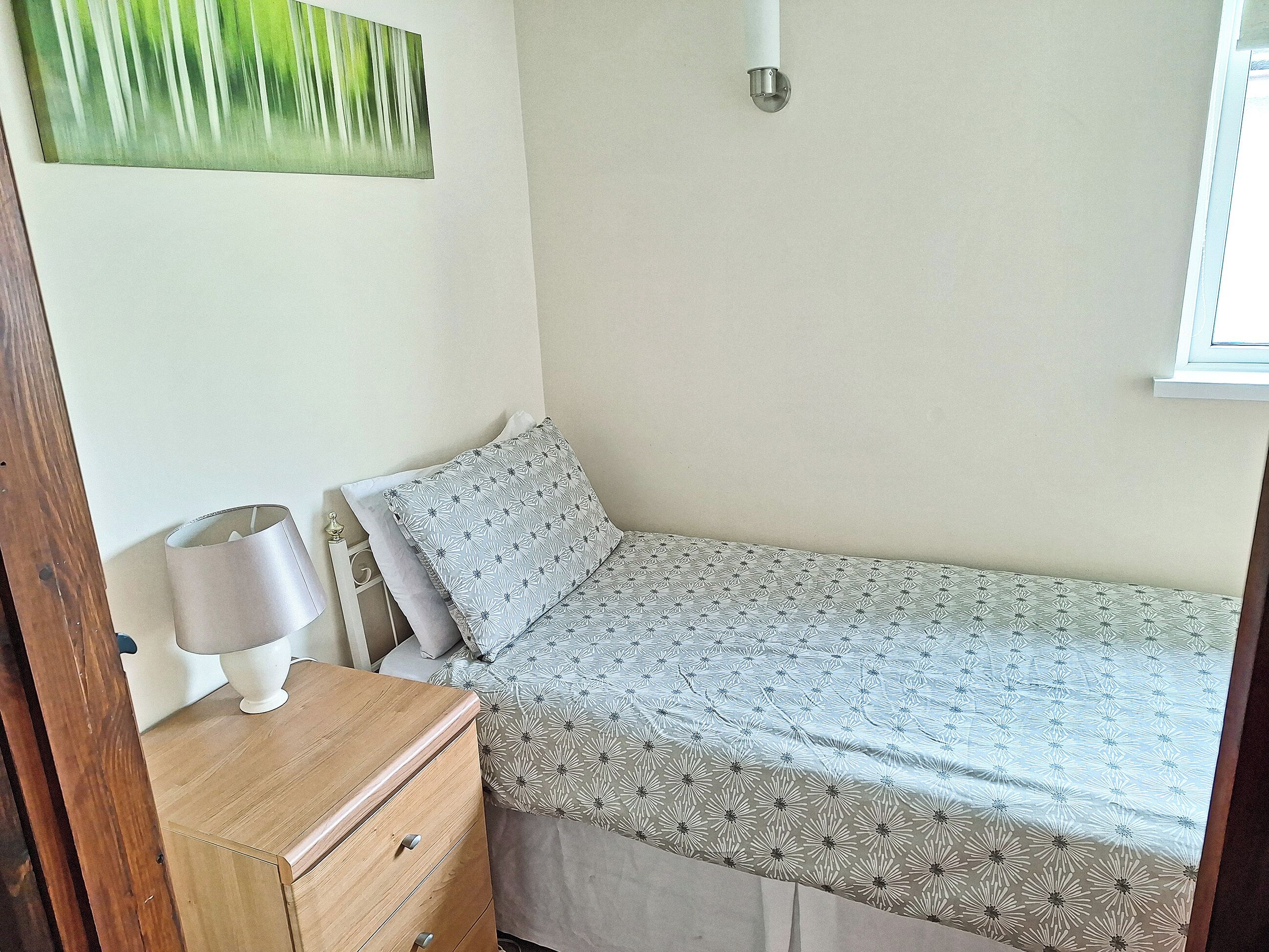 Una camera piccola con un letto singolo, un comò in legno, una lampada e una coperta a motivi.