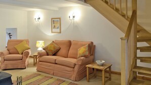 Ein Wohnzimmer mit einer Couch, einem Sessel und einem kleinen Tisch mit einer Schale. Rechts befindet sich eine Treppe und eine Wand mit zwei Wandleuchten und einem gerahmten Bild.