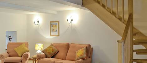 Ein Wohnzimmer mit einer Couch, einem Sessel und einem kleinen Tisch mit einer Schale. Rechts befindet sich eine Treppe und eine Wand mit zwei Wandleuchten und einem gerahmten Bild.