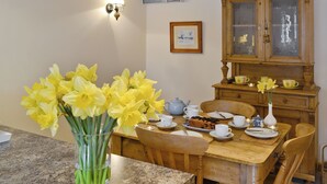 Ein Essbereich mit einem Holztisch, der für vier Personen gedeckt ist, einer Vase mit gelben Blumen und einem Holzschrank mit Glastüren.