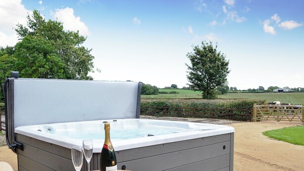 Een hottub met een fles champagne en glazen op een houten tafel, buiten met uitzicht op een boerderij en bomen.