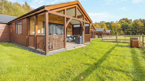 A wooden cabin with a covered porch, a fenced yard, and a view of the surrounding trees. - Humphrey-Basil Lodge-UKC2176 (Norton Disney, near Lincoln)