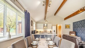 A dining area with a long table set for six, a kitchen in the background, and a living area with a sofa and a blue patterned wall. - Humphrey-Basil Lodge-UKC2176 (Norton Disney, near Lincoln)