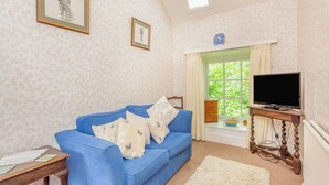 A living room with a blue sofa, a television on a wooden stand, and a window with curtains. - 1 bedroom accommodation in Lyth, near Wick (Lyth, near Wick)