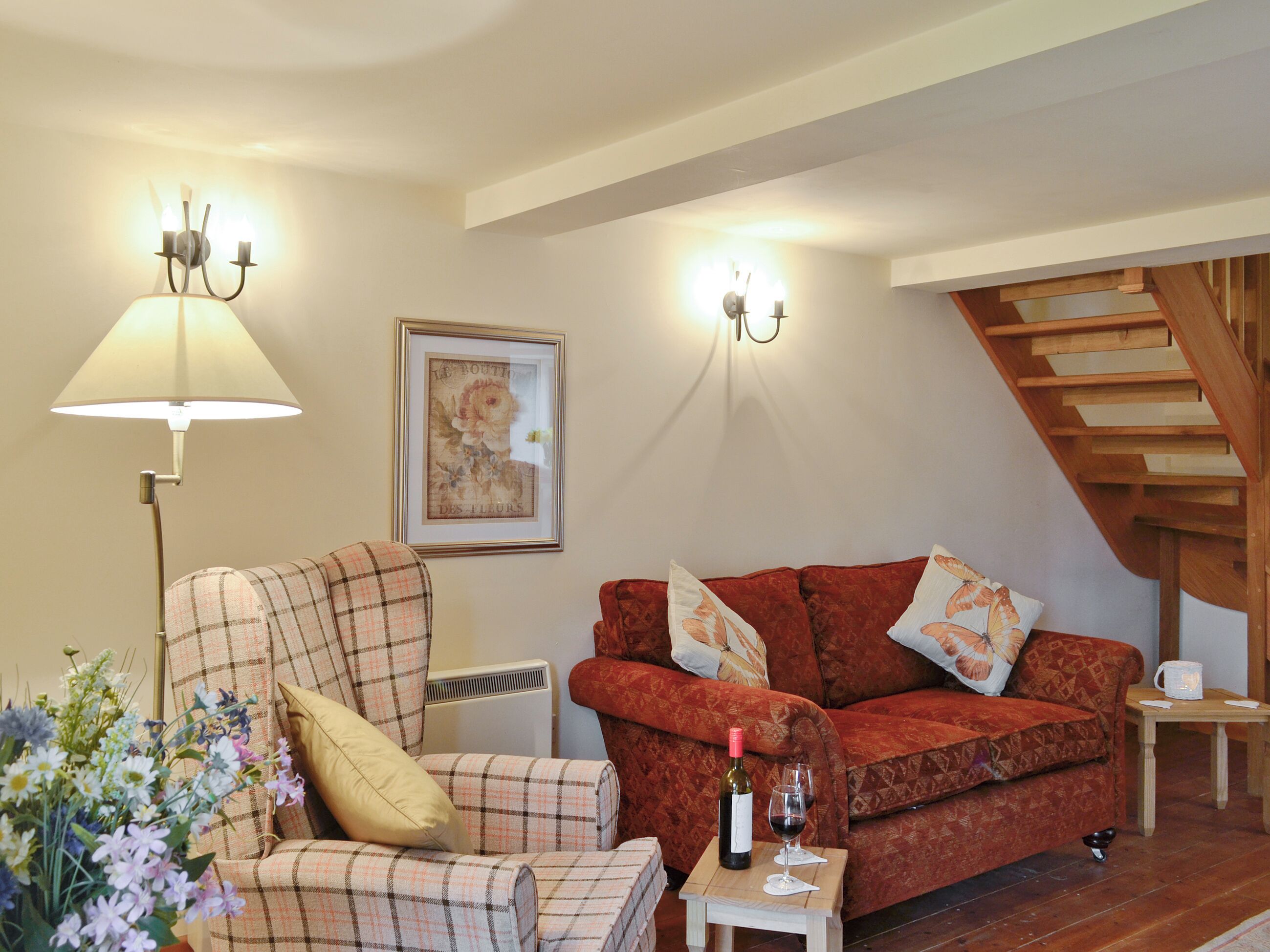 A cozy living room with a plaid armchair, a red sofa, a small table with a bottle of wine, and a framed picture on the wall.