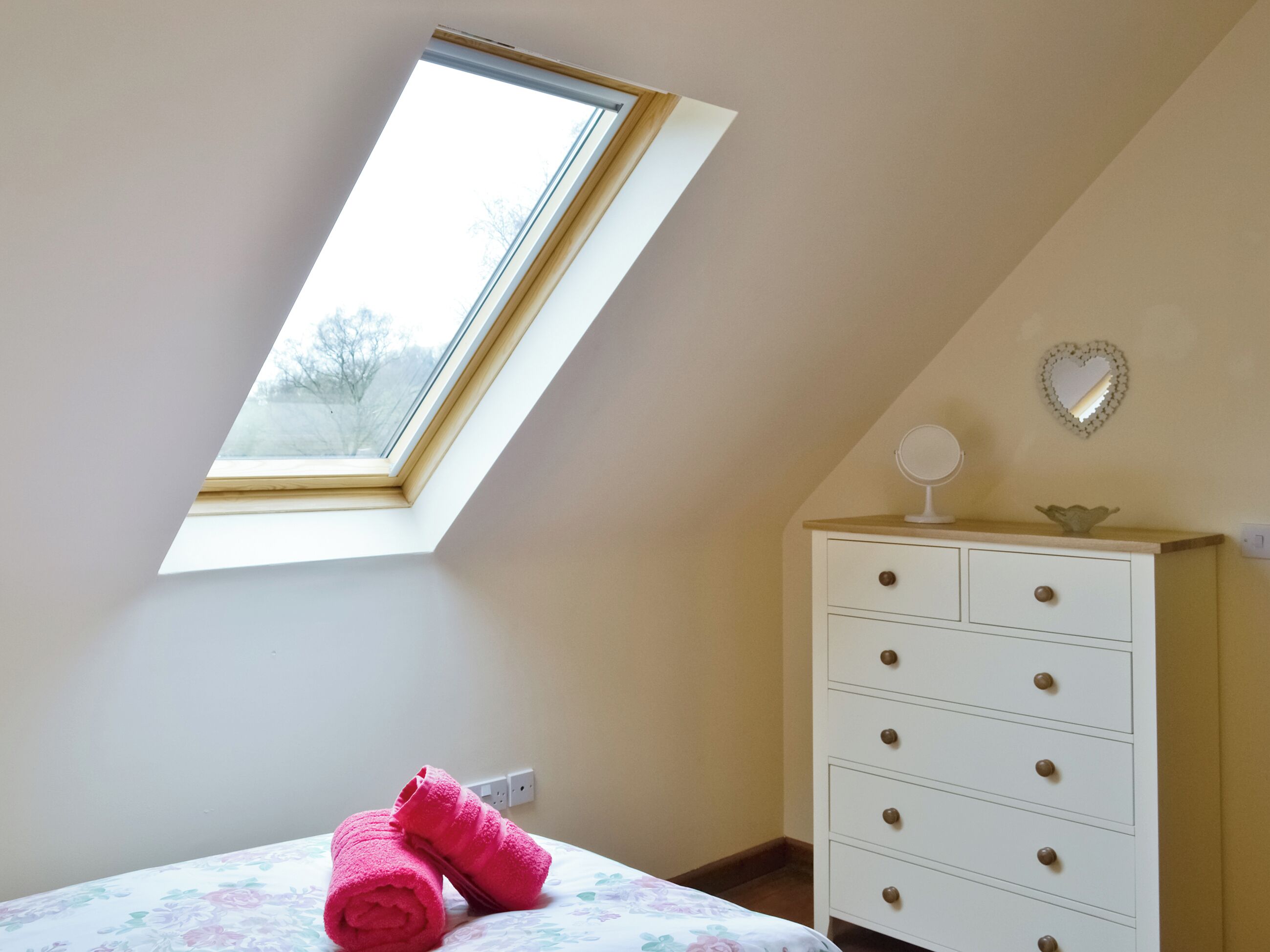 A bedroom with a skylight, a bed with floral bedding, a white dresser, and a heart-shaped mirror on the wall.