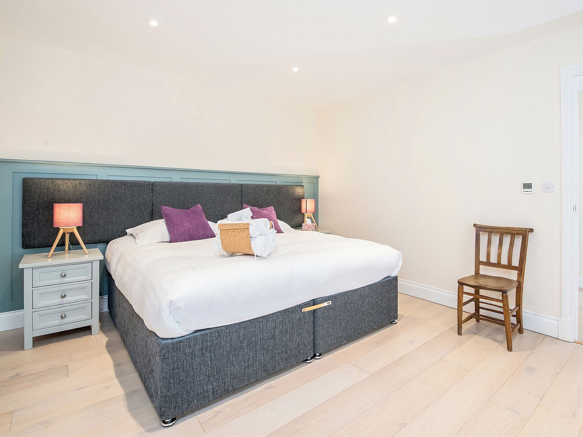 A neatly made bed with a grey headboard, two bedside tables with lamps, a wooden chair, and a small bedside drawer.