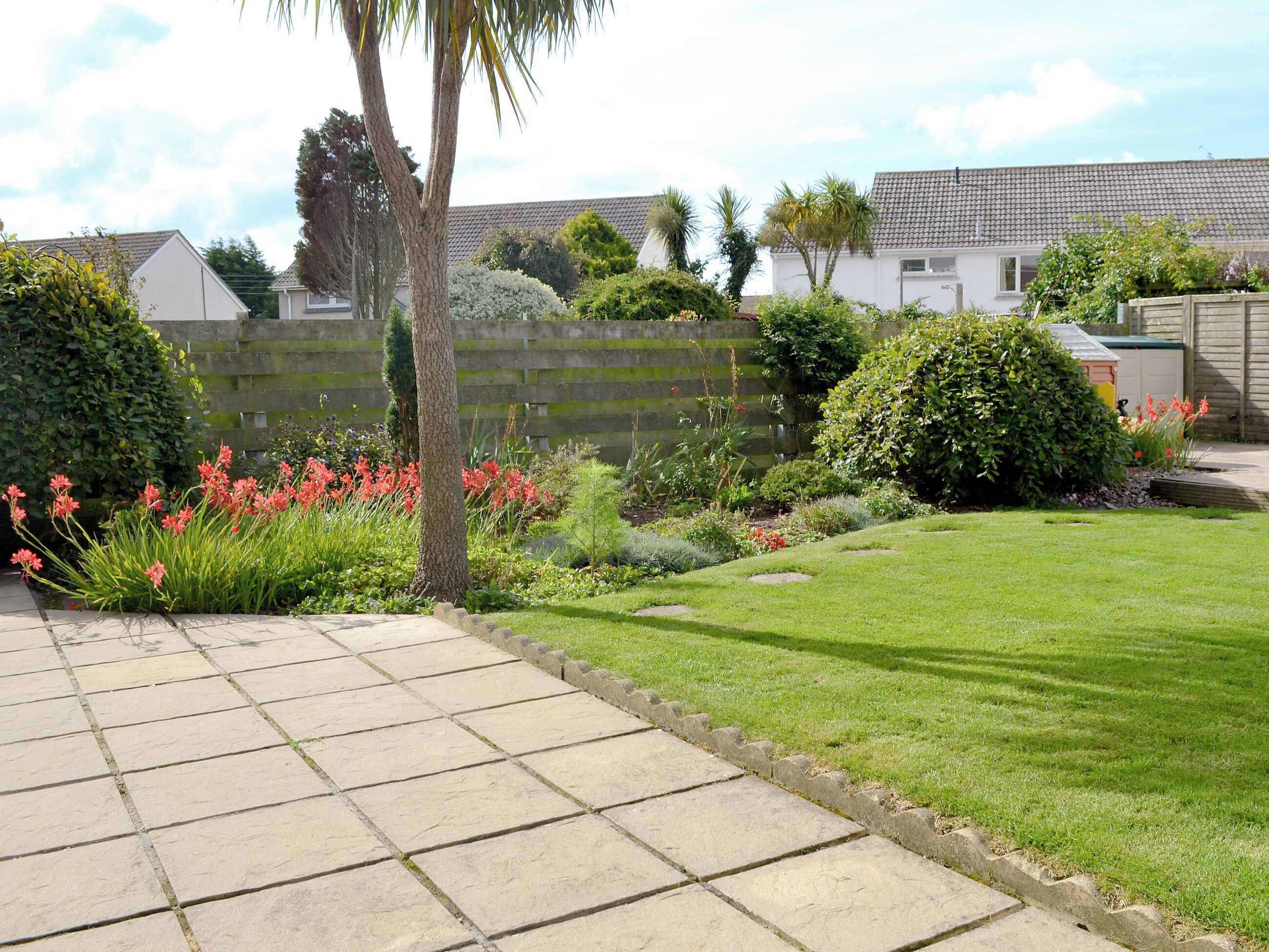 A residential garden with a paved pathway, lush greenery, and flowering plants.