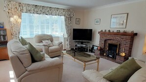 Ein Wohnzimmer mit einer Couch, einem Sessel und einem Couchtisch. Im Hintergrund ist ein Kamin, ein Fernseher und ein Bücherregal zu sehen.