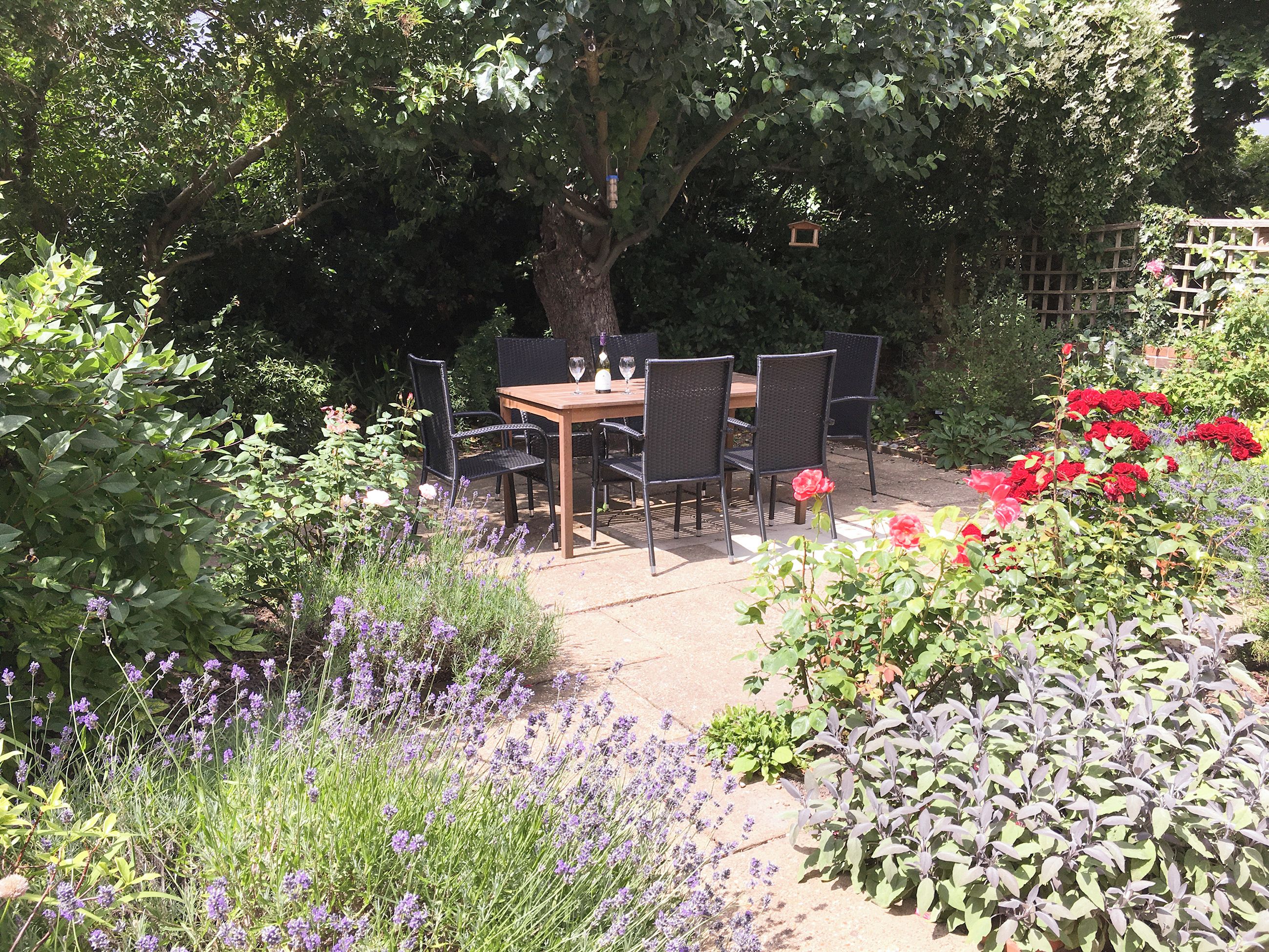 An outdoor garden patio with a wooden table and chairs, surrounded by lush greenery and flowering plants.