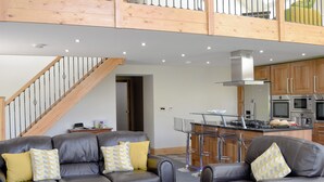 Una sala moderna con un sofá, sillones, una mesa de centro de vidrio y una zona de cocina con electrodomésticos.