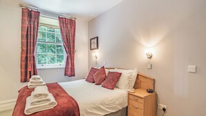 A neatly made bed with a red blanket and white pillows, a wooden nightstand with a lamp, a window with curtains, and a wall-mounted light fixture. - Apartment 14, Hazelwood Hall (Silverdale, near Arnside)