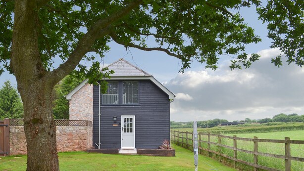 A two-story house with a stone foundation and dark siding, a front door, and a small front yard with a wooden fence. - Swift Cottage - UKC3435 (East Ogwell, near Newton Abbot)