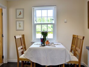 A dining area with a table set for two, a window with a view of greenery, and two framed pictures on the wall.