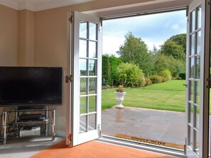 An open sliding glass door leading to a lawn with a potted plant, a television on a stand, and a doormat.