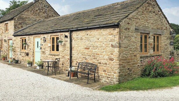 A stone cottage with a gravel driveway, a small patio with a table and chairs, and a flower bed with pink flowers.