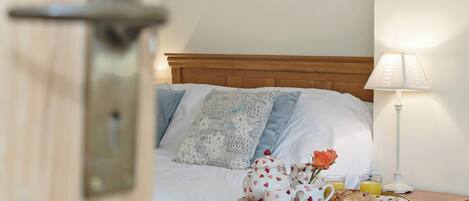 Una cama bien hecha con cabecera de madera, una mesita de noche con lámpara y una bandeja con alimentos para el desayuno.