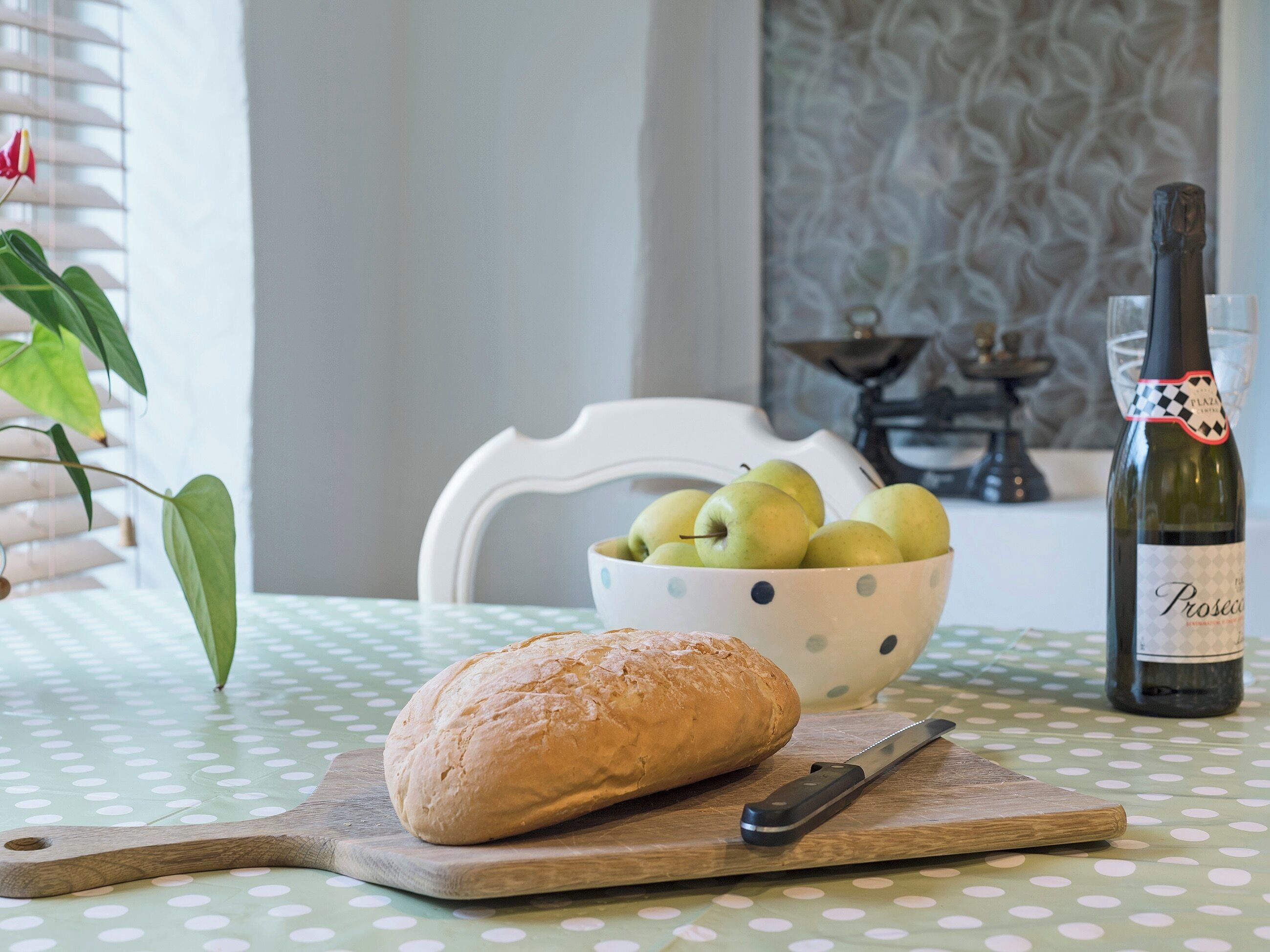 A table with a loaf of bread, a knife, a bowl of green apples, and a bottle of wine.