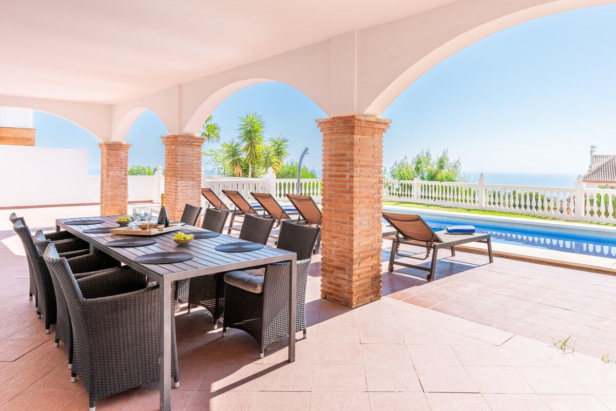 Un patio exterior con mesa y sillas, piscina y vista al mar.