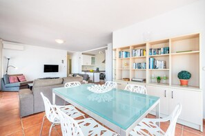 A modern living room with a glass-top dining table, white chairs, a grey sofa, and a bookshelf with books and decorative items. - Casa Pamela | Spacious Family Villa with Two Private Pools, One Heated, & Sea Views in Benalmadena Pueblo (Benalmádena)