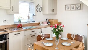 A modern kitchen with white cabinetry, a wooden dining table set for four, and a clock on the wall. - 2 bedroom accommodation in Beadnell (Beadnell)
