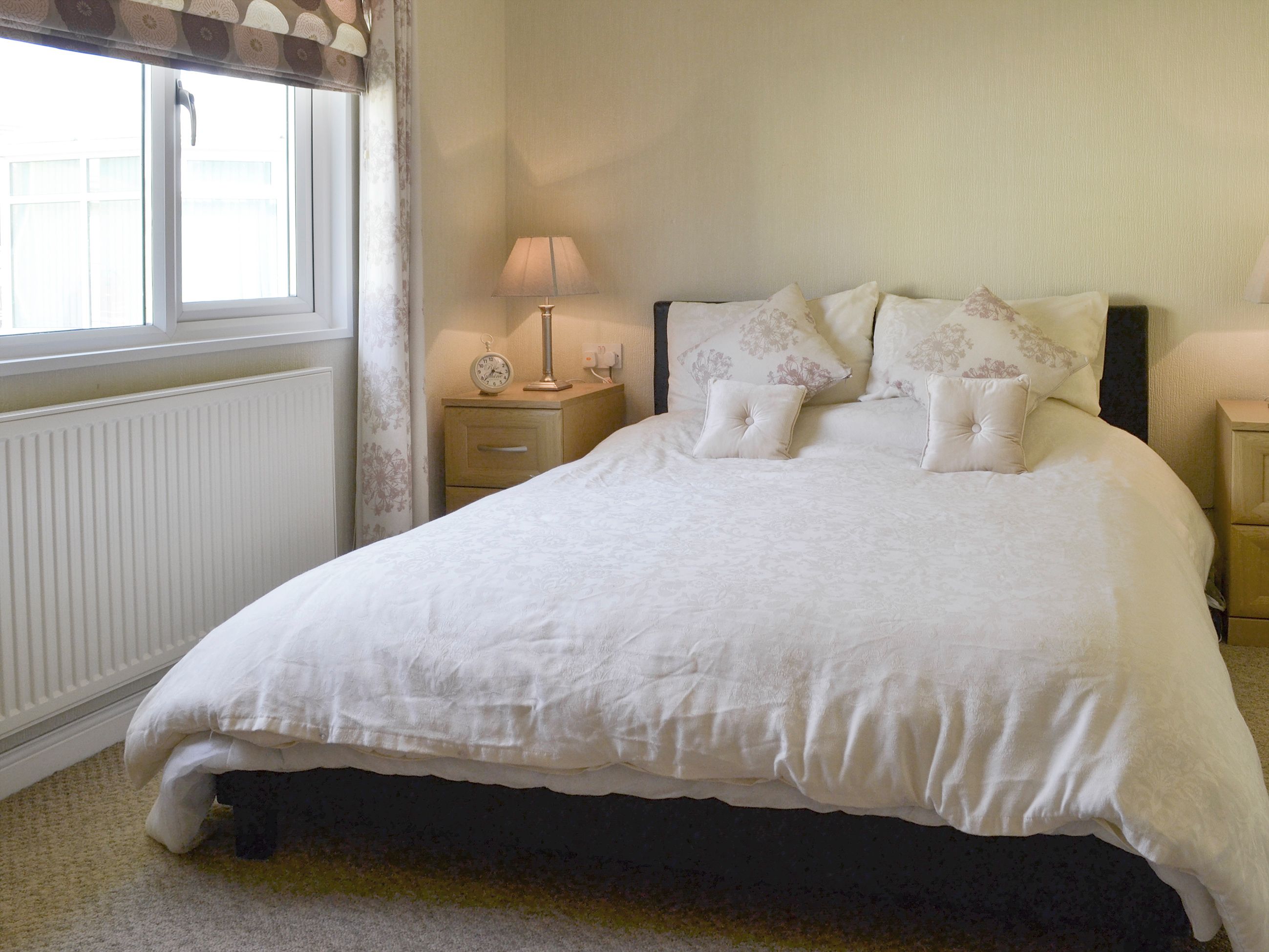 A neatly made bed with a white duvet and floral pillows, a bedside table with a lamp and a clock, and a window with curtains.