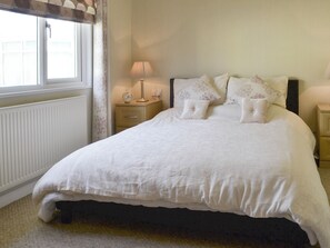 A neatly made bed with a white duvet and floral pillows, a bedside table with a lamp and a clock, and a window with curtains.