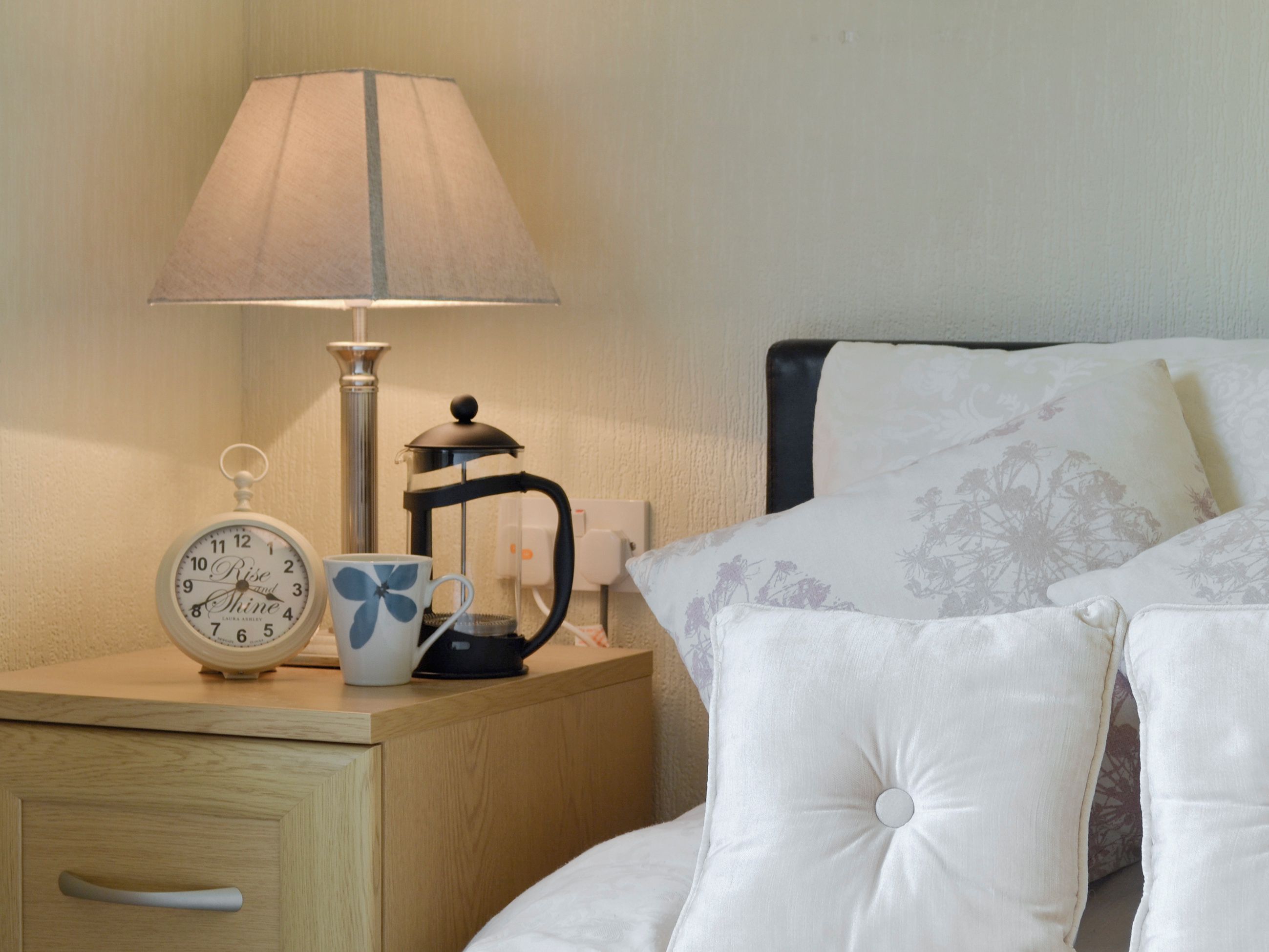 A neatly arranged bedroom with a bedside table featuring a clock, a lamp, a coffee maker, and a decorative mug.