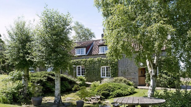 Een huis met een stenen gevel, een pannendak en een tuin met een houten tafel en stoelen.