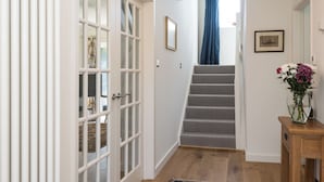 Un pasillo con suelo de madera, una escalera, una puerta con paneles de vidrio y un jarrón con flores sobre una mesa de madera.