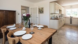 Un comedor con una mesa de madera preparada para cuatro personas, platos blancos y copas de vino. Hay un jarrón con flores sobre la mesa.
