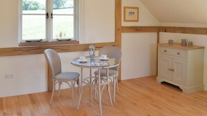 A cozy room with a round table, two chairs, a window with a valance, a wooden ceiling, and a small cabinet with two drawers.