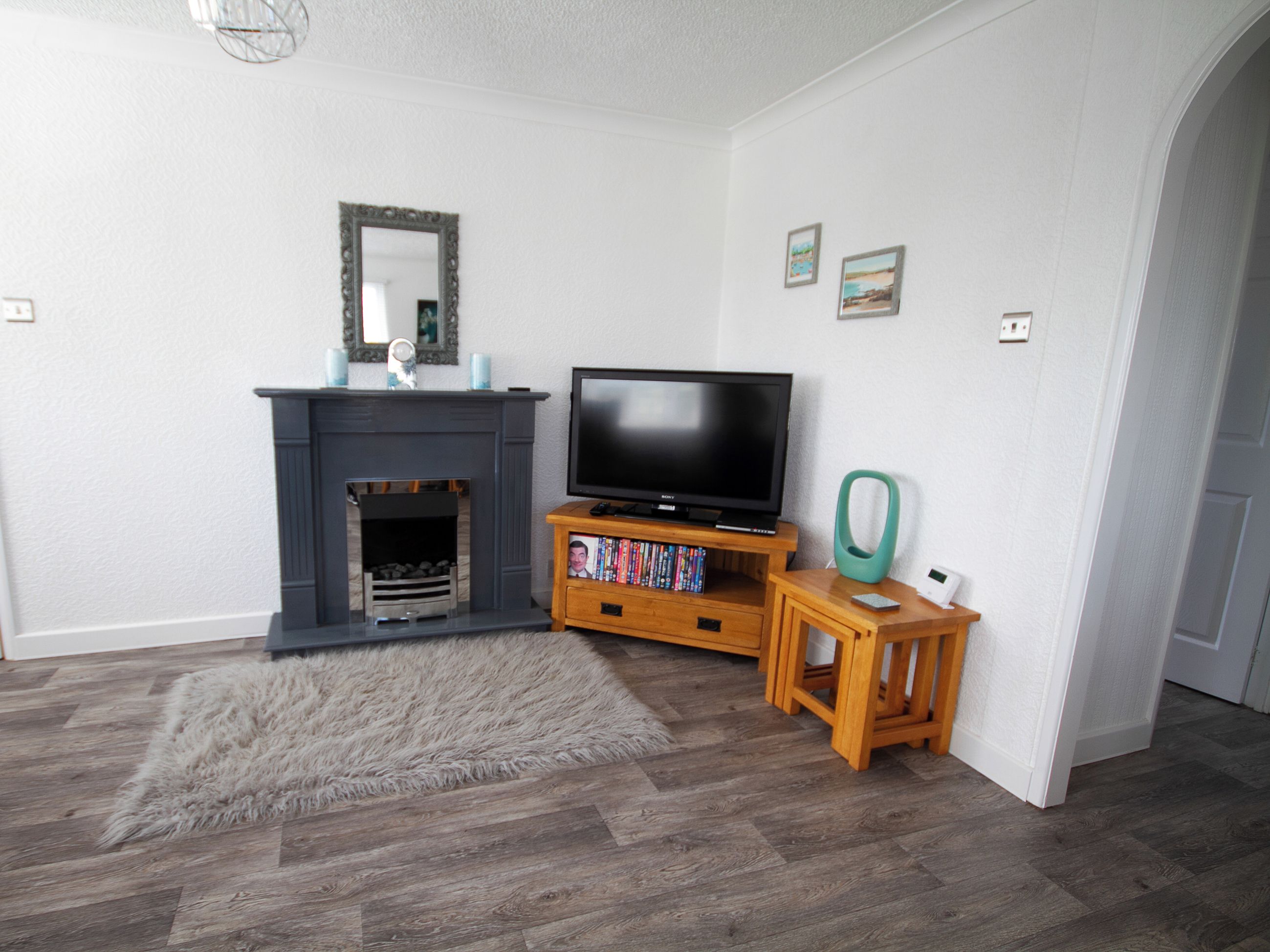 A living room with a fireplace, a TV on a wooden stand, a small table with a green chair, and a fluffy rug.