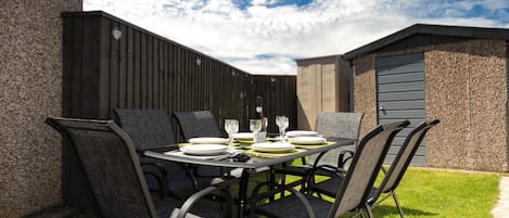 Un área de comedor al aire libre con una mesa preparada para cuatro personas, rodeada de sillas, con un cerco de madera y un edificio al fondo.