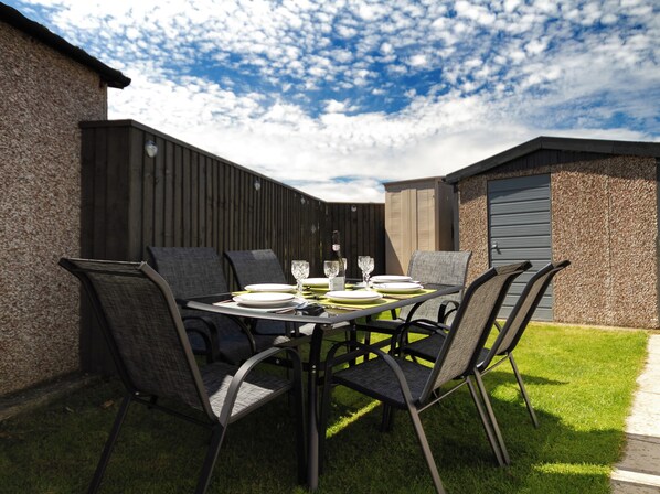 Un espace extérieur aménagé pour les repas, avec une table dressée pour quatre personnes, entourée de chaises, le tout adossé à une clôture en bois et à un bâtiment.