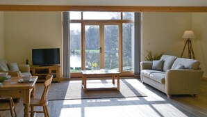 A spacious living room with a wooden floor, a sofa, a dining table with chairs, a TV, and a large window with a view of trees.