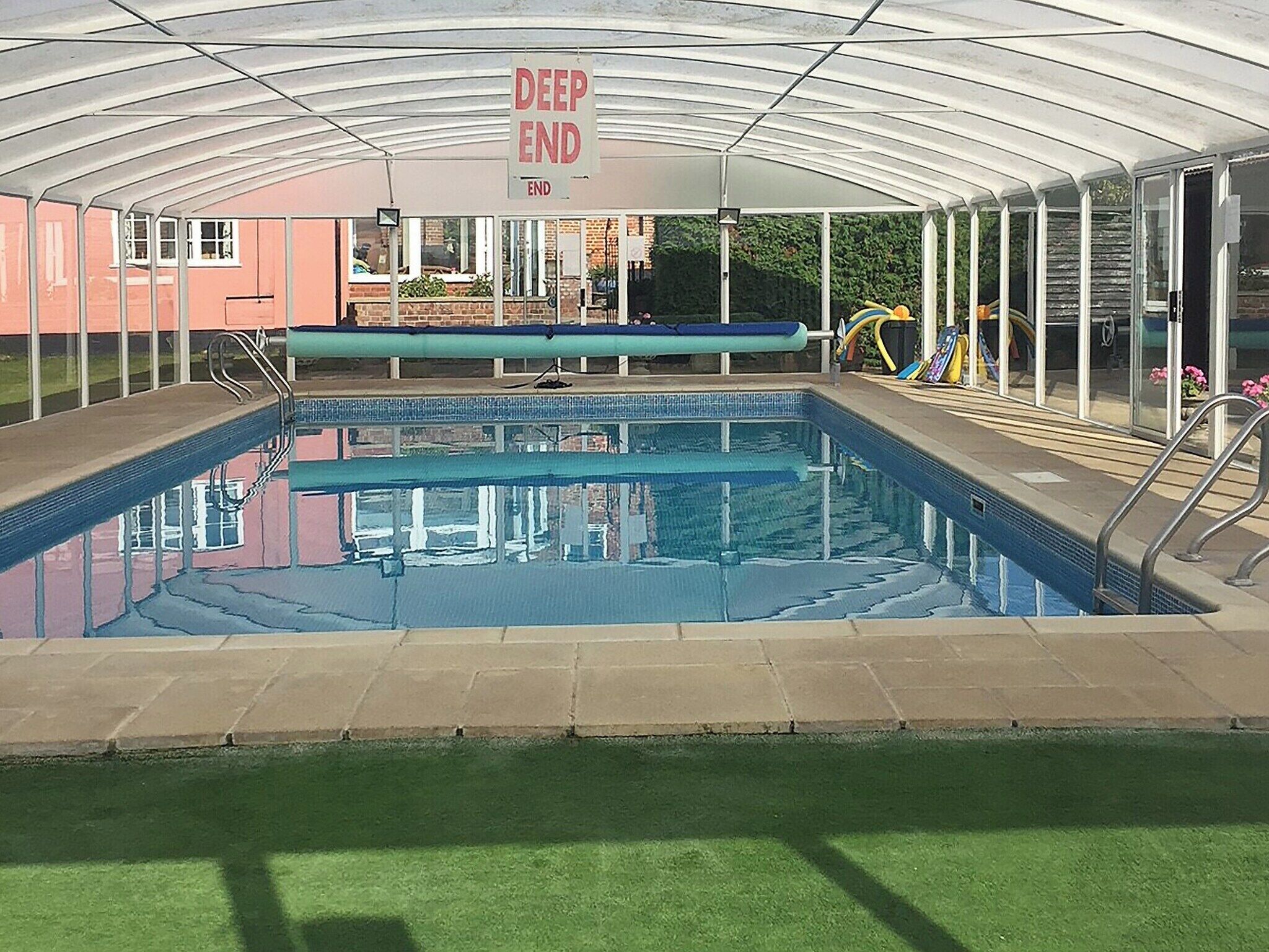 A covered swimming pool with a 'DEEP END' sign, surrounded by a glass enclosure and a grassy area.