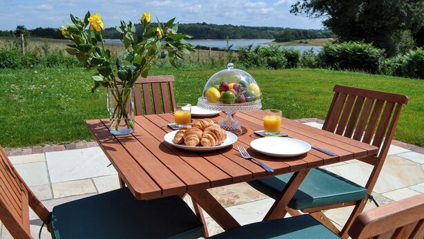 A wooden table set outdoors with a view of a lake, a fruit bowl, croissants, and a glass of orange juice. - 2 bedroom accommodation in Wadhurst, near Tunbridge Wells (Wadhurst, near Tunbridge Wells)