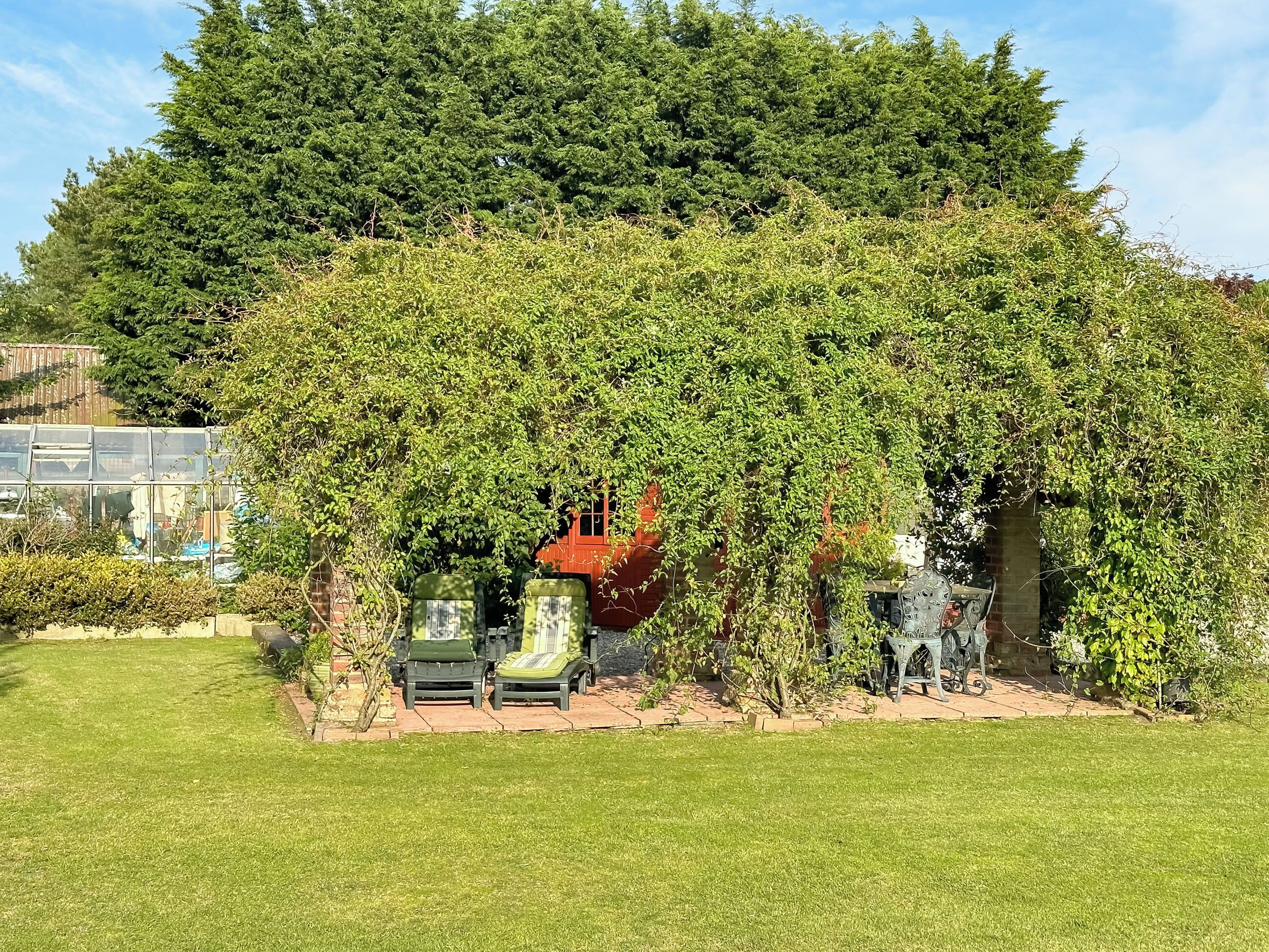 Un jardín con un pérgola cubierta de hiedra, una estructura roja y dos sillas verdes.