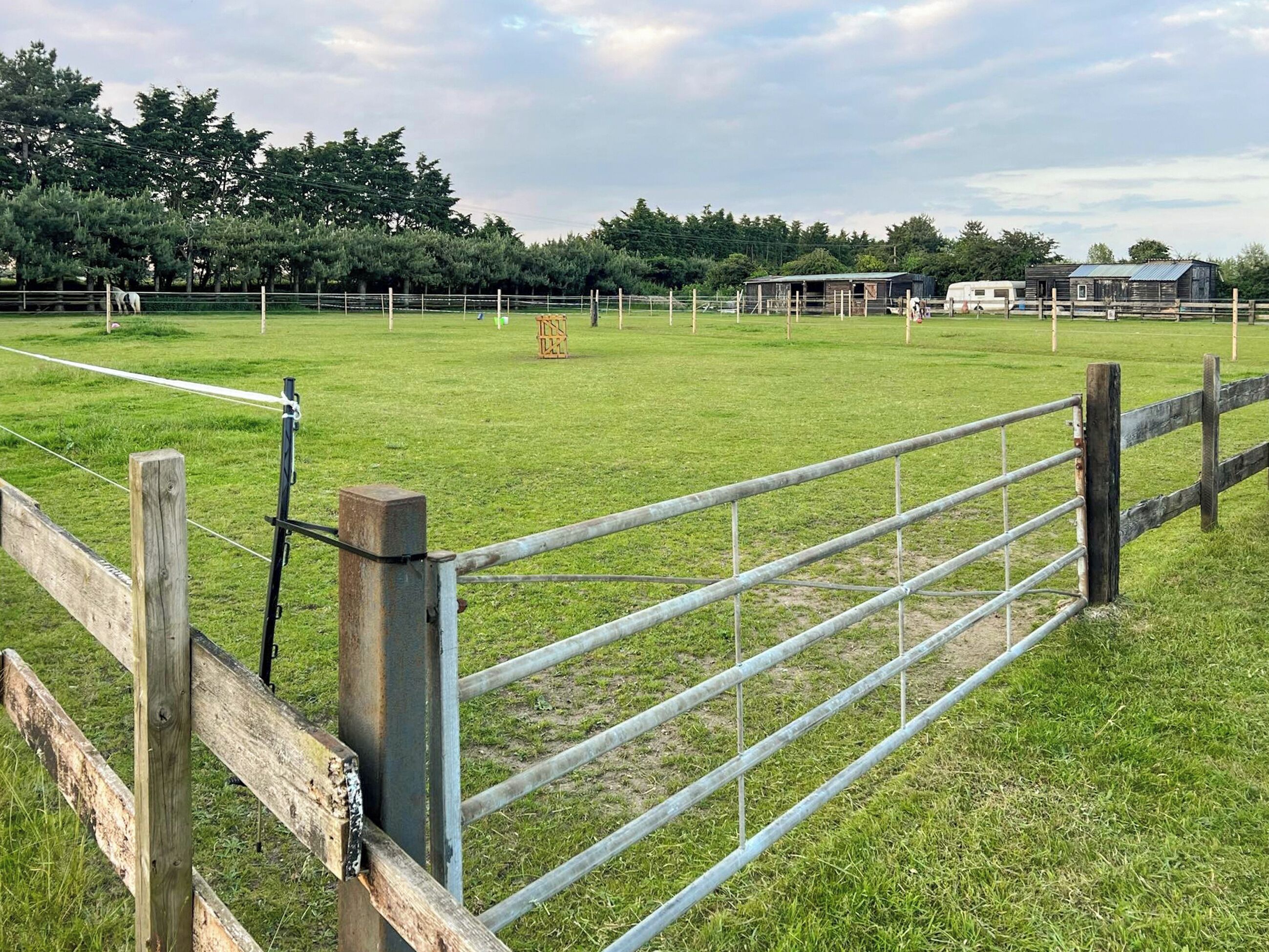 A fenced pasture with a wooden post and metal gate, surrounded by trees and a building in the background.