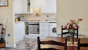 Een compacte keuken met witte kastjes, een vaatwasser en een kookplaat. Er is een eettafel met stoelen en een bloevas.