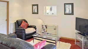 Ein Wohnzimmer mit schwarzem Ledersofa, einem roten und weißen Kissen, einem Glastisch, einem weißen Bücherregal und einem Flachbildfernseher.