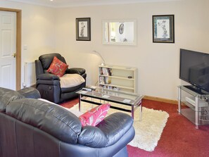 A living room with a black leather sofa, a red and white patterned throw pillow, a glass coffee table, a white bookshelf, and a flat-screen TV. - 1 bedroom accommodation in Polperro, near Looe (Polperro, near Looe)
