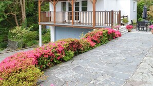 Ein weißes Gebäude mit einem hölzernen Balkon, umgeben von rosafarbenen Blumen und Pflastersteinen.